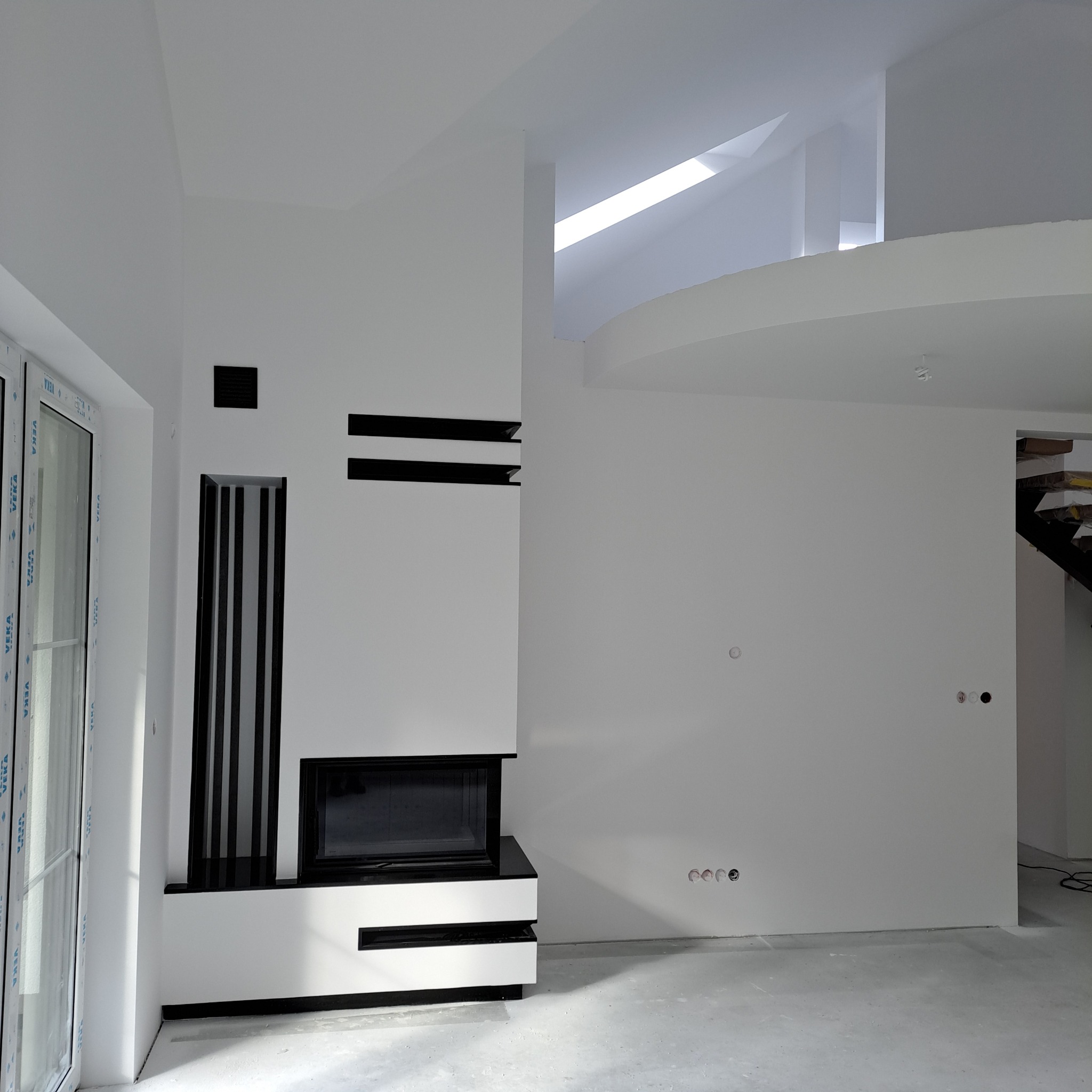Nowoczesny salon z białymi ścianami i kominkiem w zabudowie z czarnymi akcentami. Widoczne okno i fragment schodów. Minimalistyczny design wnętrza.