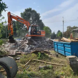 Pomarańczowa koparka Doosan kruszy gruz ceglany, z boku widoczny niebieski kontener, w tle zieleń drzew i błękitne niebo.