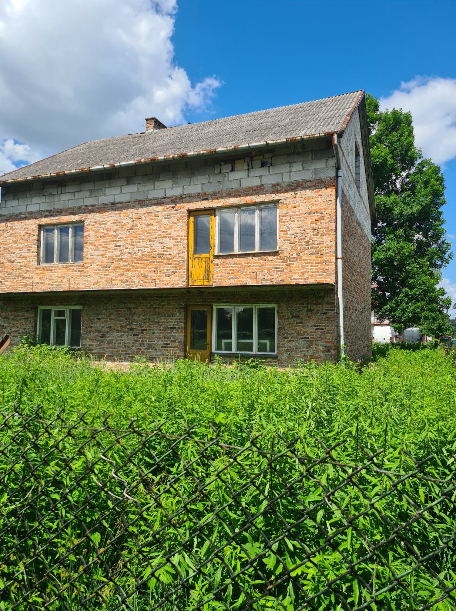 Dwukondygnacyjny budynek z czerwonej cegły z żółtymi drzwiami, częściowo wykończony, otoczony bujną zielenią i ogrodzeniem z siatki, widok w słoneczny dzień.