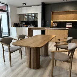 Jasny drewniany stół z owalnym blatem i żłobionymi nogami w nowoczesnej kuchni z białymi i grafitowymi szafkami oraz drewnianymi frontami, uzupełniony szarymi krzesłami z drewnianymi ramami.