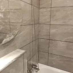 Wykończona łazienka z beżowymi płytkami imitującymi marmur, biała wanna z chromowaną armaturą i okrągłe lustro w tle.