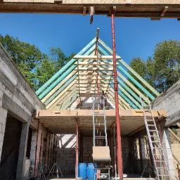 Konstrukcja dachu w trakcie budowy domu murowanego, widoczne drewniane krokwie pomalowane impregnatem, ściany z betonu komórkowego, podpory stalowe, drabina i podnośnik.