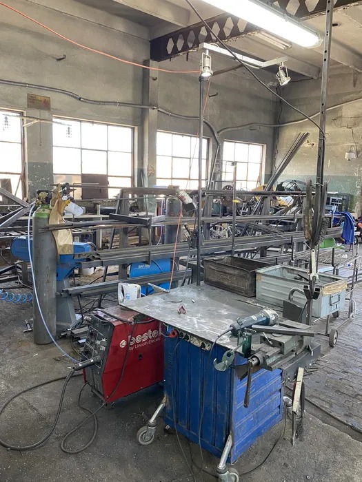 Warsztat spawalniczy: czerwona spawarka Bestel, butla z gazem, metalowy stół warsztatowy na kółkach z zamontowanym imadłem i szlifierką kątową, w tle konstrukcje z metalowych profili.