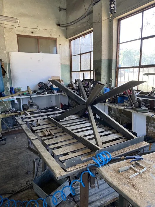 Metalowa konstrukcja w kształcie gwiazdy na stole warsztatowym w hali, widoczne narzędzia i niebieski przewód pneumatyczny, w tle okna i tablica.