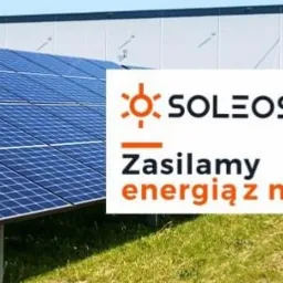 Panel słoneczny na trawie przed budynkiem, z logo firmy SOLEOS i hasłem 'Zasilamy energią z natury'.