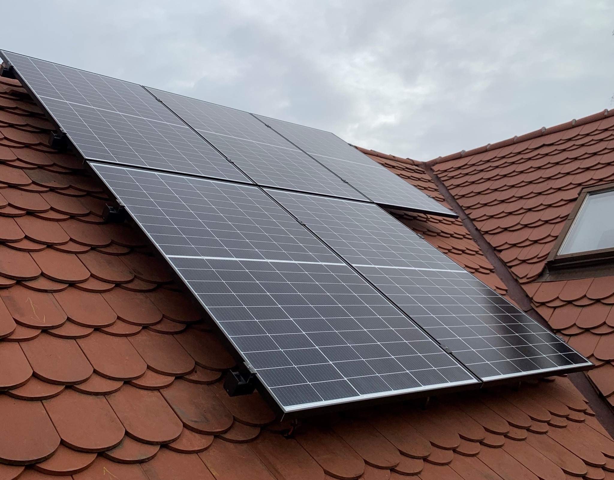 Część instalacji paneli słonecznych na dachu z czerwonej dachówki karpiówki, widok z dołu w pochmurny dzień.