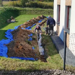 Dwaj mężczyźni instalują rury w wykopie na trawniku obok domu jednorodzinnego, zabezpieczonym niebieską folią. Jeden pracuje w wykopie, drugi z taczką na krawędzi.