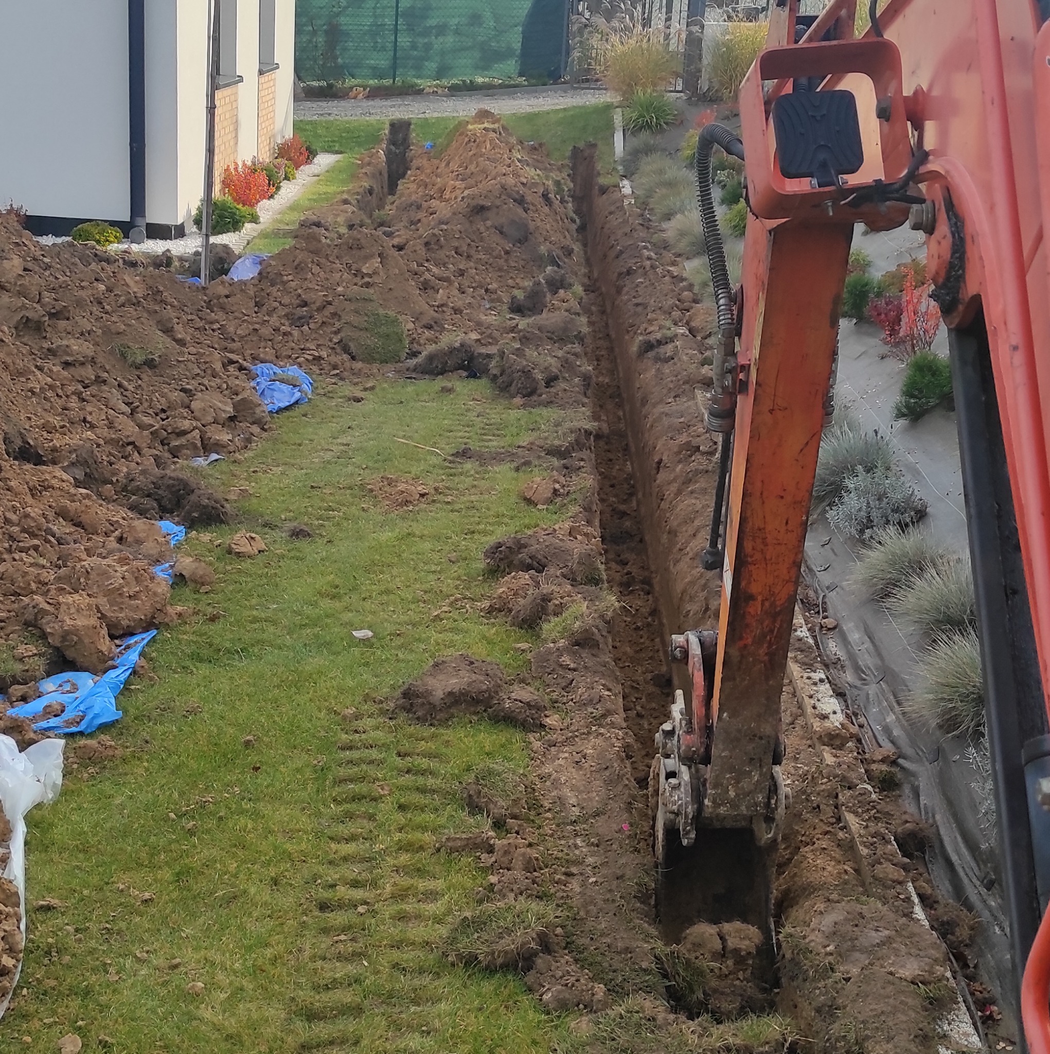 Pomarańczowa koparka wykonuje wąski wykop w ziemi na trawniku, widoczne ślady gąsienic, obok wykopu pryzma ziemi zabezpieczona niebieską folią, w tle budynek mieszkalny i roślinność.