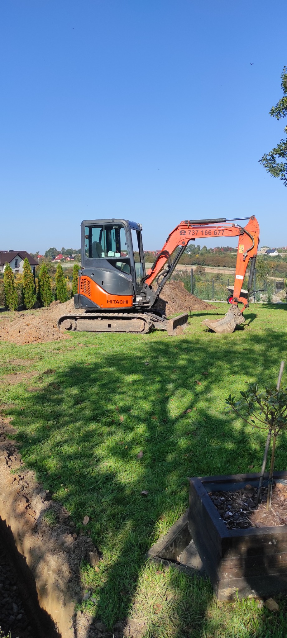 Pomarańczowo-szara mini koparka Hitachi na trawniku w słoneczny dzień, obok świeżo wykopany rów, w tle domy i drzewa.