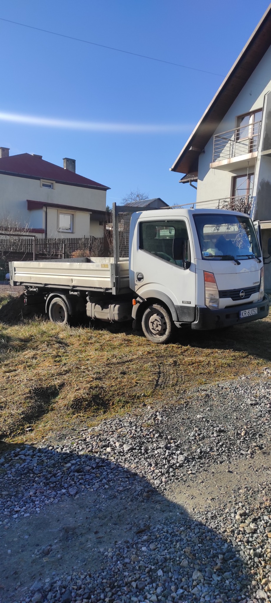 Biała ciężarówka Nissan Cabstar z otwartą skrzynią ładunkową, zaparkowana na nierównym terenie z kamieniami i trawą, w tle domy jednorodzinne pod błękitnym niebem.