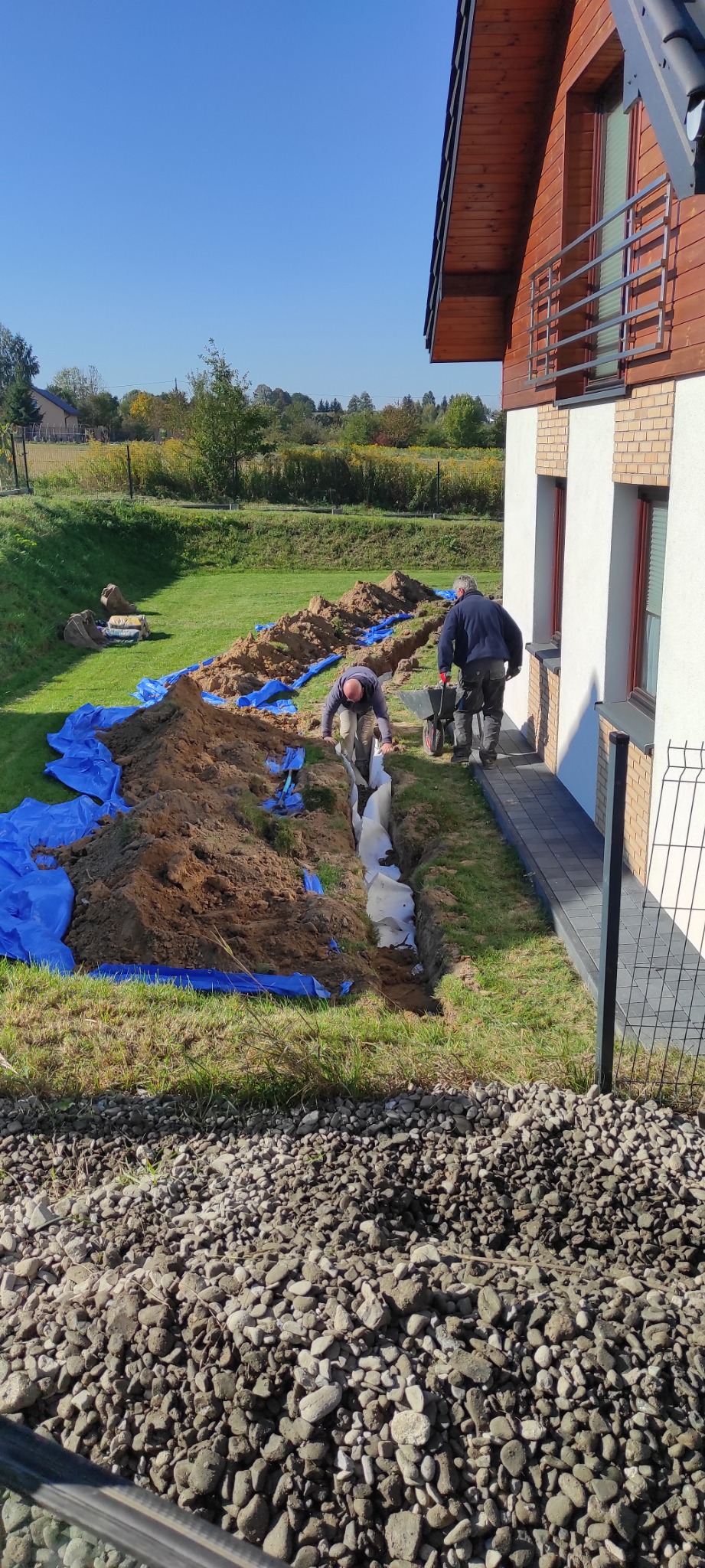 Dwaj mężczyźni instalują rury w wykopie na trawniku obok domu jednorodzinnego, zabezpieczonym niebieską folią. Jeden pracuje w wykopie, drugi z taczką na krawędzi.
