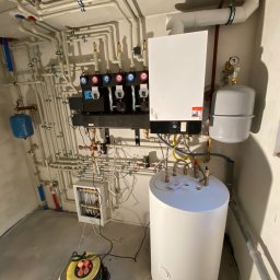 Alpineclima Michał Zagrabski - Węzeł cieplny w kotłowni: biały zasobnik ciepłej wody, szary zbiornik przeponowy, biały kocioł gazowy Viessmann z czarnym panelem sterowania, rozdzielacz z czterema obiegami grzewczymi...