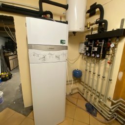 Alpineclima Michał Zagrabski - Biały piec gazowy Vaillant z podłączonym zasobnikiem ciepłej wody i rurami instalacji centralnego ogrzewania w kotłowni z beżowymi płytkami na podłodze i widocznymi narzędziami w tle.