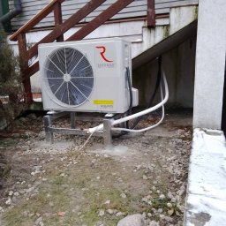 Alpineclima Michał Zagrabski - Zewnętrzna jednostka pompy ciepła Rotenso Aquami Series zamontowana na metalowej konstrukcji, widoczne rury prowadzące do budynku, tło: drewniane schody i elewacja budynku.