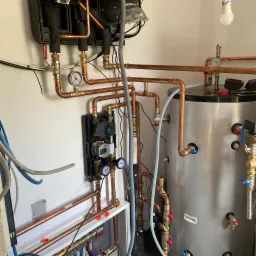 Instalacja grzewcza z miedzianymi rurami, zbiornikiem i czarną skrzynką rozdzielczą na białej ścianie. Widoczne manometry, zawory i kable. Żarówka zwisa z sufitu.