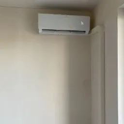 Nowy, biały klimatyzator ścienny, jeszcze w folii ochronnej, zamontowany w jasnym pomieszczeniu z kremowymi ścianami i białymi framugami drzwi.