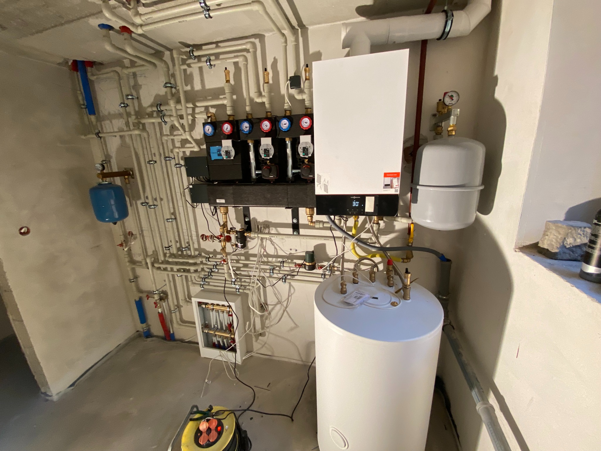 Węzeł cieplny w kotłowni: biały zasobnik ciepłej wody, szary zbiornik przeponowy, biały kocioł gazowy Viessmann z czarnym panelem sterowania, rozdzielacz z czterema obiegami grzewczymi...