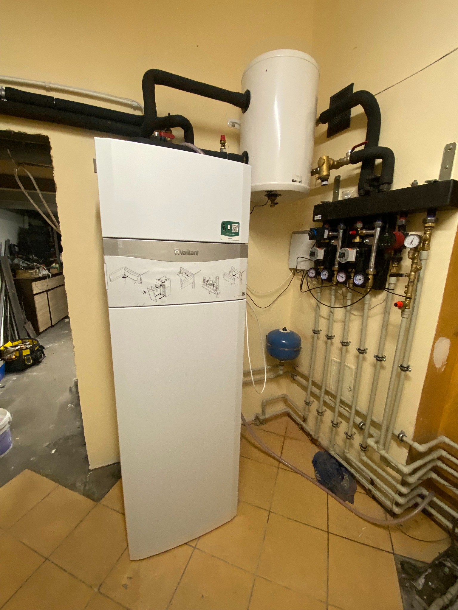 Biały piec gazowy Vaillant z podłączonym zasobnikiem ciepłej wody i rurami instalacji centralnego ogrzewania w kotłowni z beżowymi płytkami na podłodze i widocznymi narzędziami w tle.