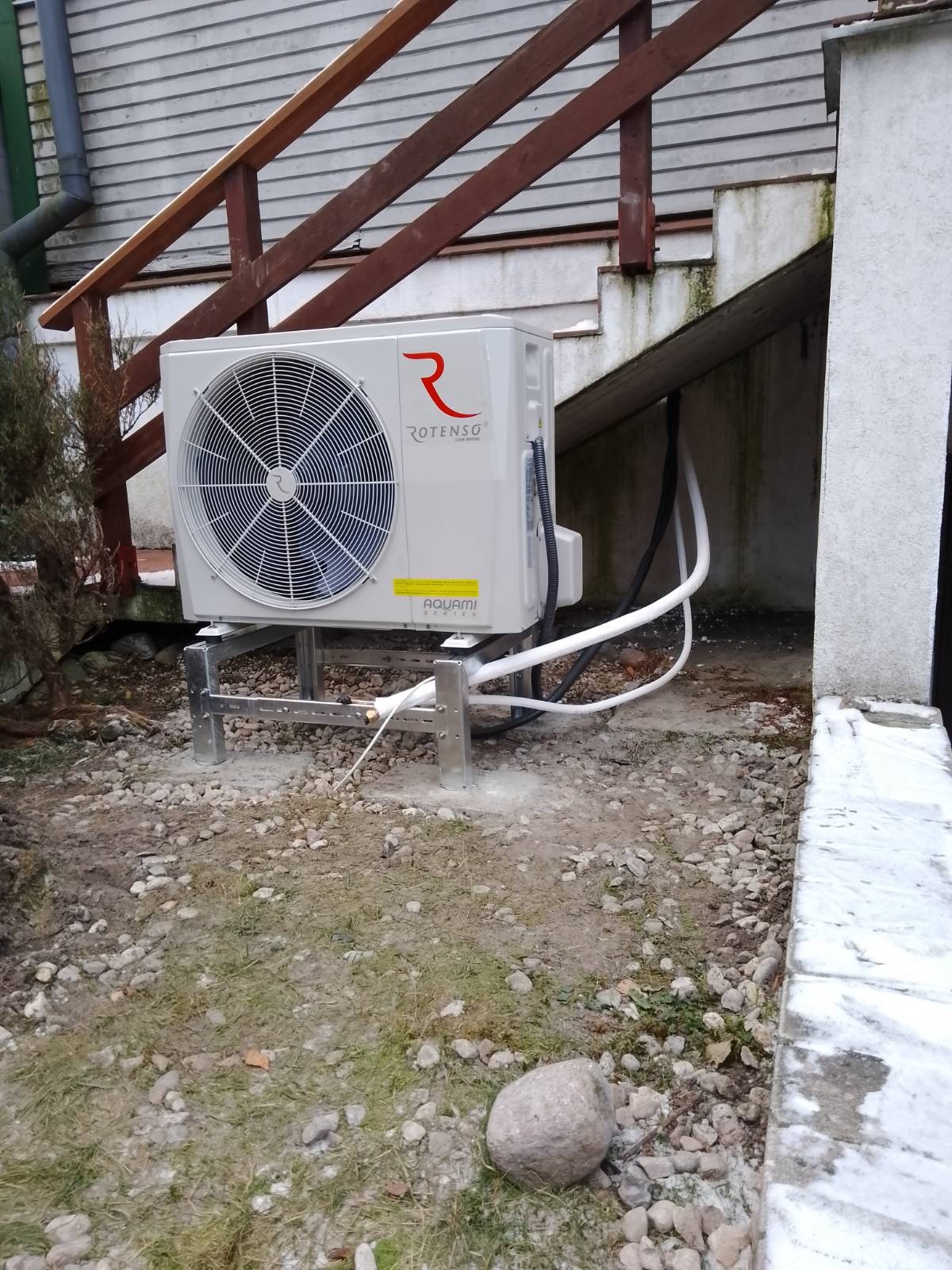 Zewnętrzna jednostka pompy ciepła Rotenso Aquami Series zamontowana na metalowej konstrukcji, widoczne rury prowadzące do budynku, tło: drewniane schody i elewacja budynku.