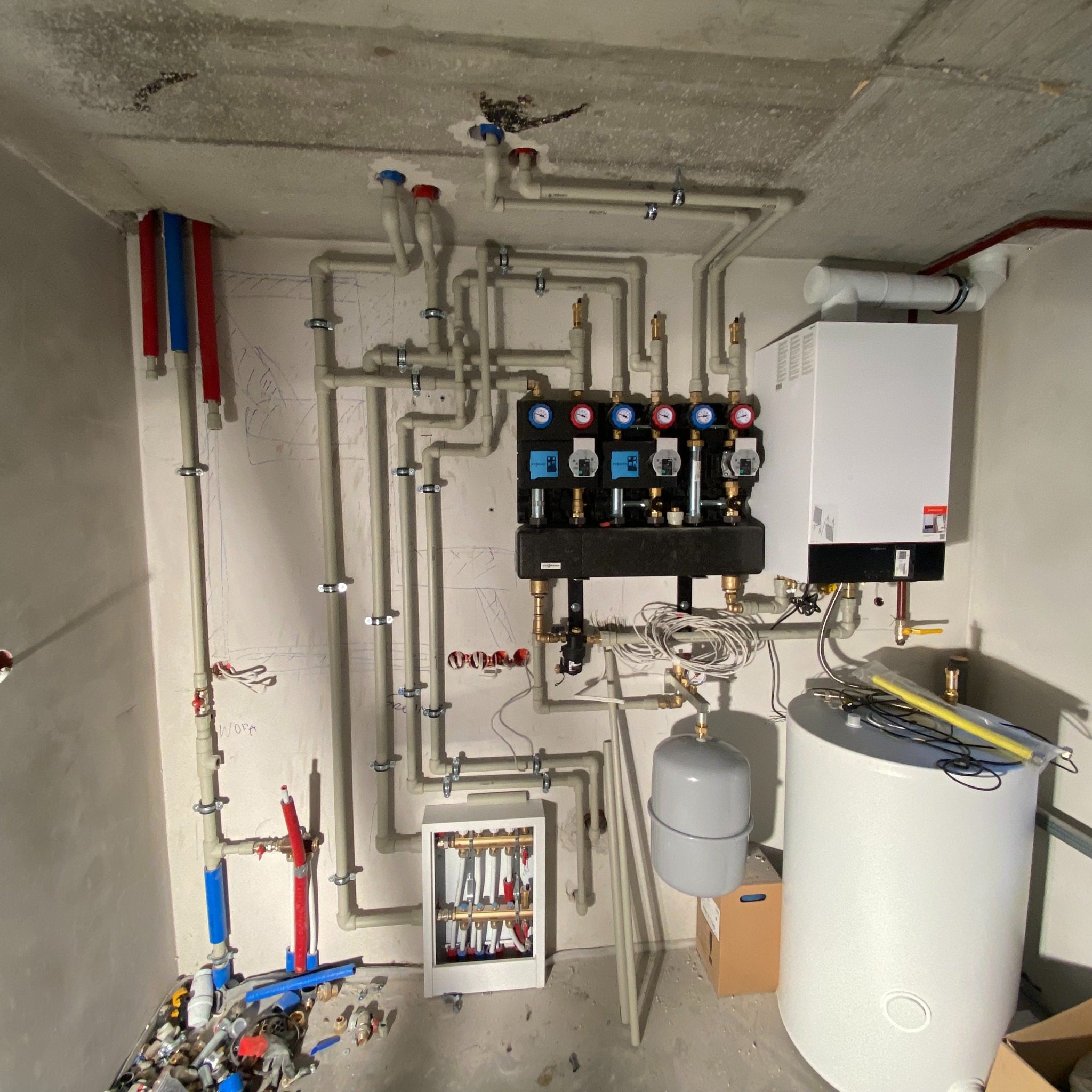 Rozbudowana instalacja grzewcza z kotłem, zbiornikiem CWU i rozdzielaczami, widoczne rury z manometrami, całość zamontowana na surowej ścianie w trakcie prac wykończeniowych.