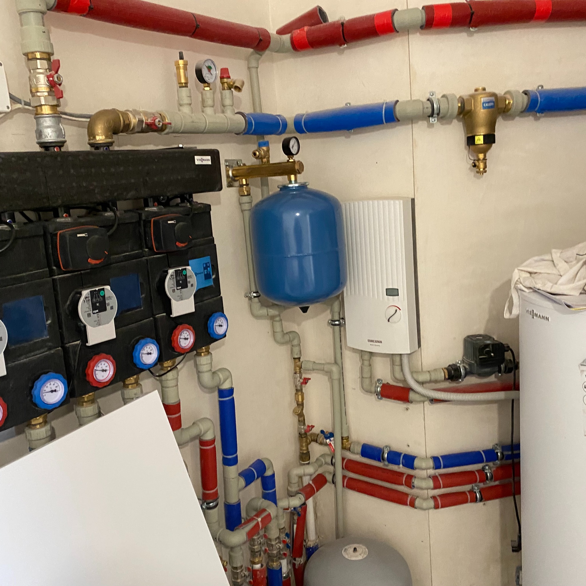 Rozbudowana instalacja grzewcza z licznymi rurami (czerwone i niebieskie), zaworami, manometrami, zbiornikiem wyrównawczym w kolorze niebieskim oraz białym zasobnikiem ciepłej wody marki Viessmann,...