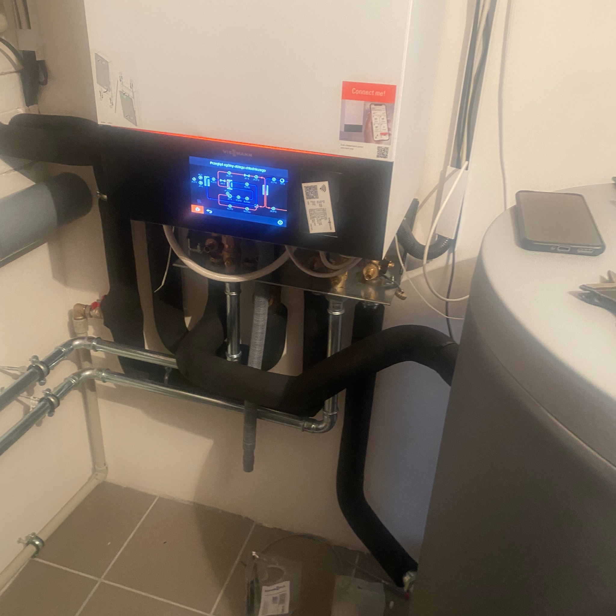 Nowoczesna instalacja grzewcza Viessmann z ekranem dotykowym, widoczne rury izolowane termicznie, w tle zasobnik wody i telefon komórkowy leżący na nim.