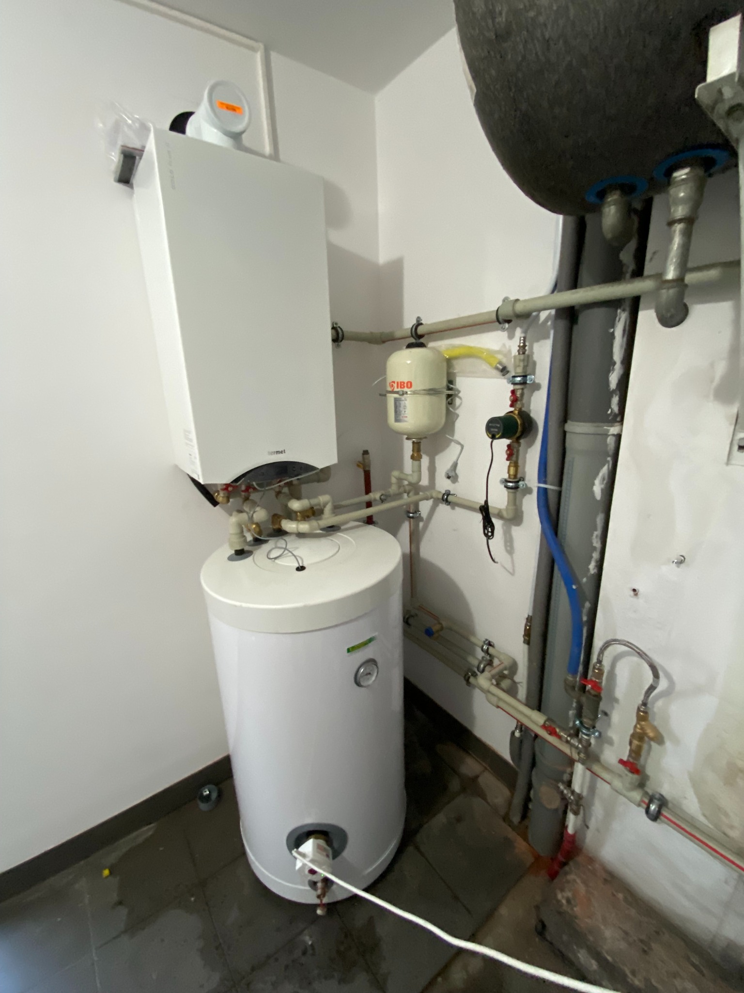 Biały, wiszący piec gazowy Termet z podłączonym zasobnikiem ciepłej wody, widoczne rury instalacyjne, naczynie przeponowe i pompa obiegowa w kotłowni.