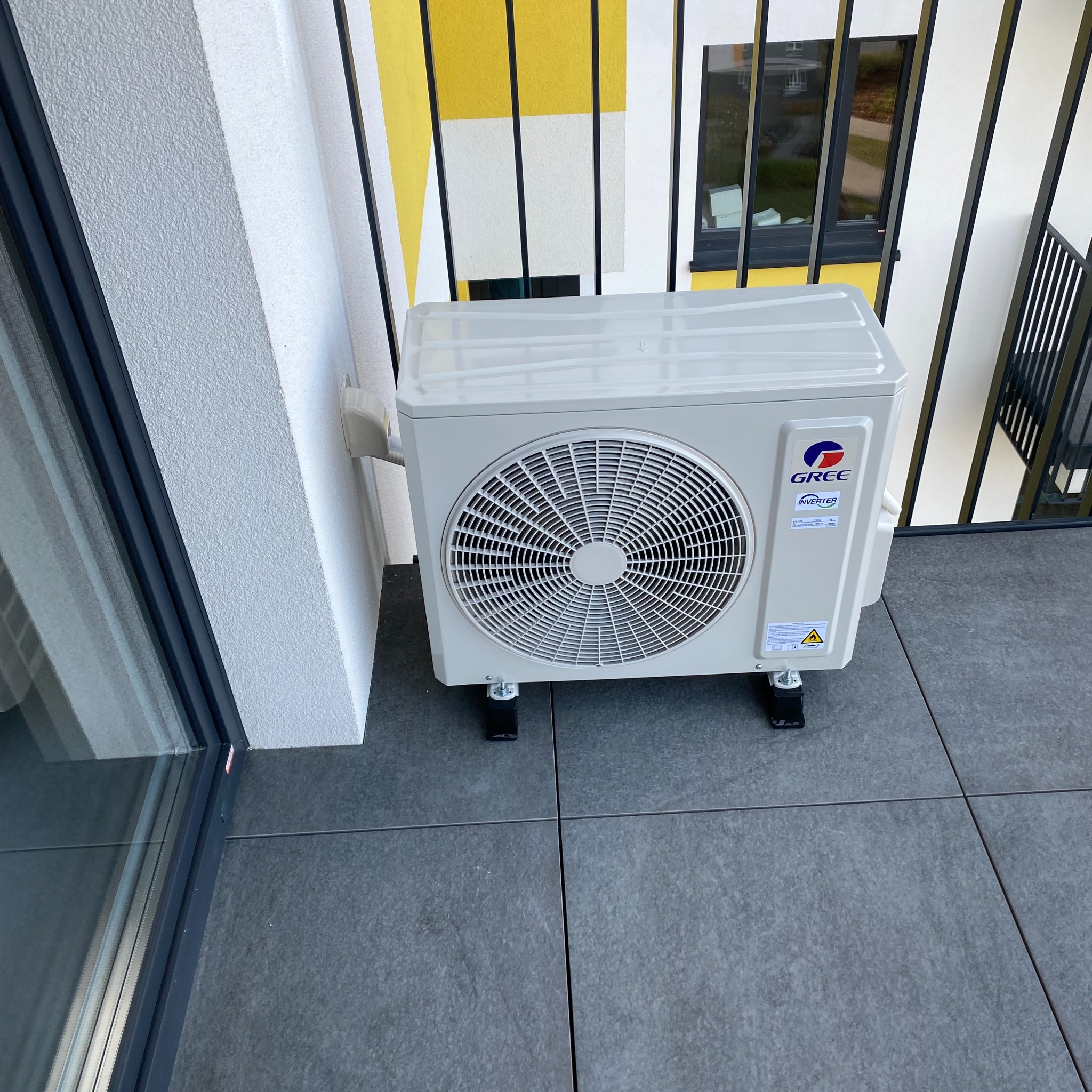 Biała jednostka zewnętrzna klimatyzatora marki Gree, zamontowana na czarnych nóżkach na balkonie z szarymi płytkami, widoczne żółto-białe elementy elewacji budynku w tle.
