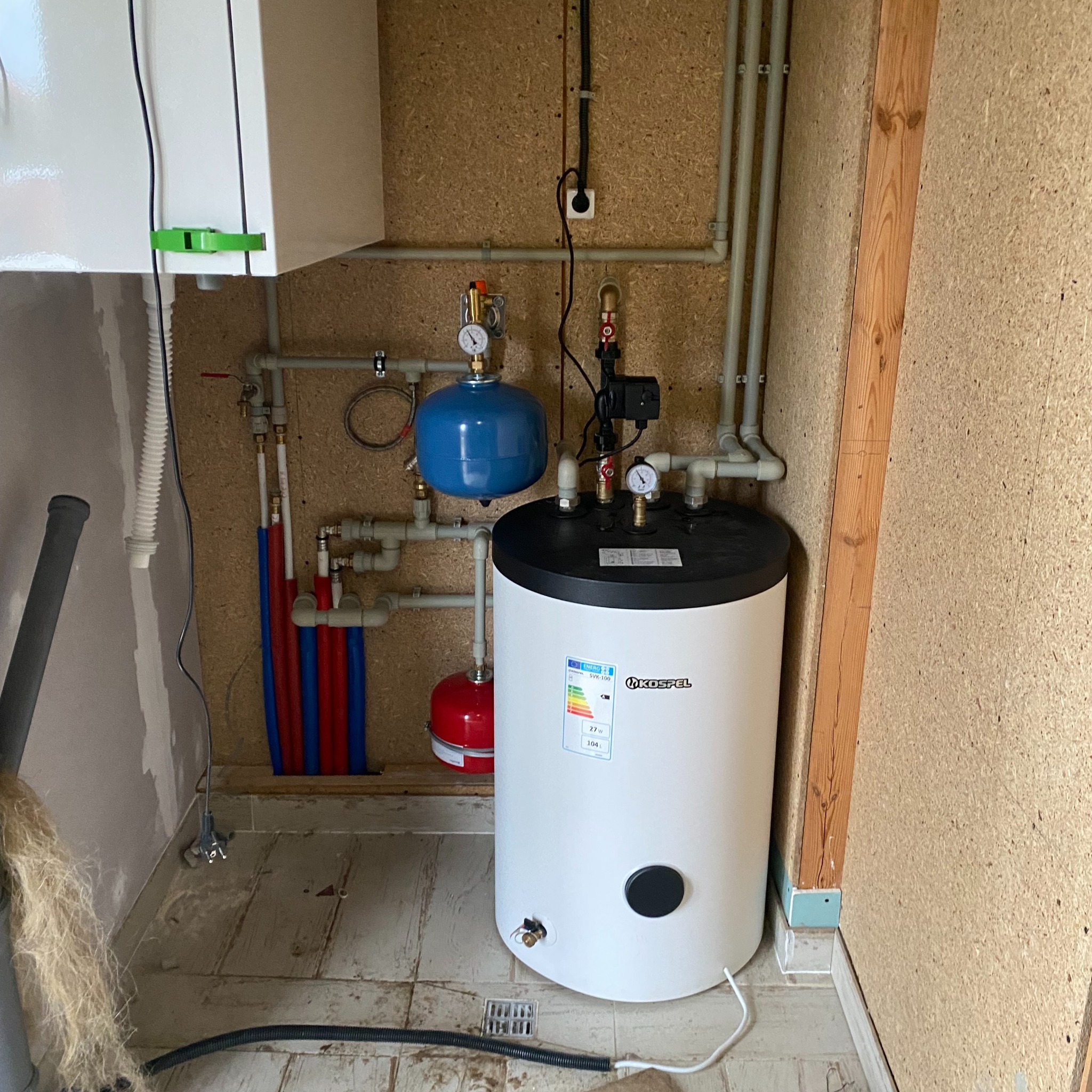 Zamontowany biały bojler Kospel z czarnym panelem, widoczne rury miedziane, plastikowe czerwone i niebieskie, oraz dwa zbiorniki wyrównawcze: niebieski i czerwony, na ścianie płyta OSB