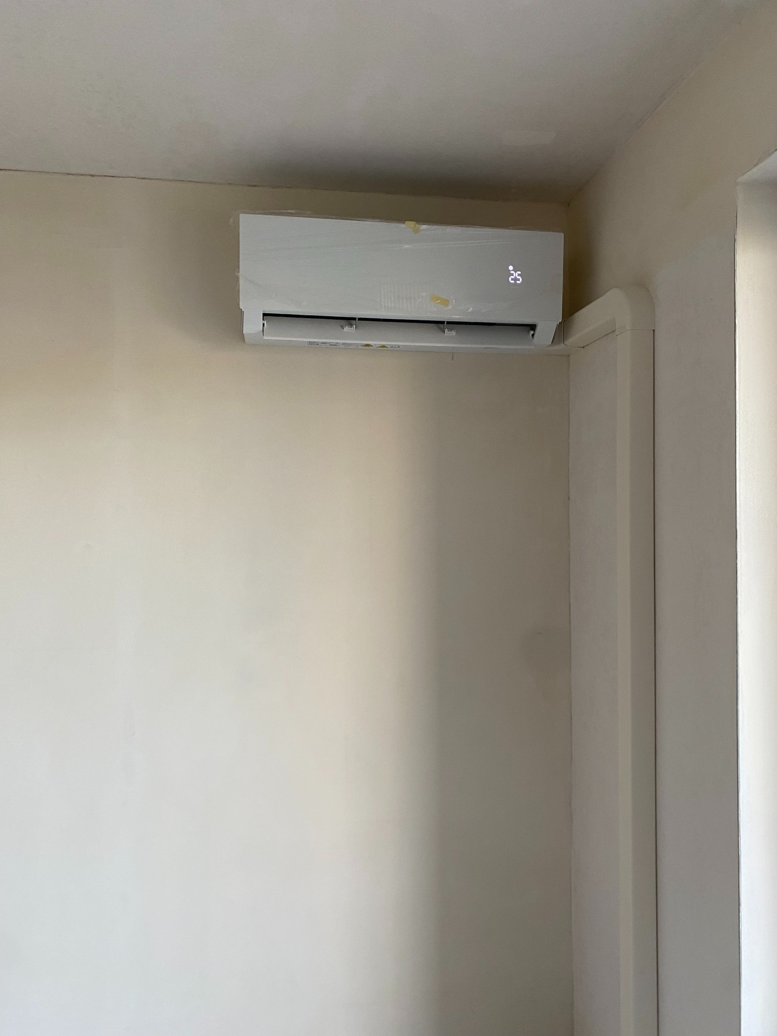 Biały klimatyzator ścienny, jeszcze w folii ochronnej, zamontowany w rogu jasnego pomieszczenia, wyświetlający temperaturę 25 stopni Celsjusza.