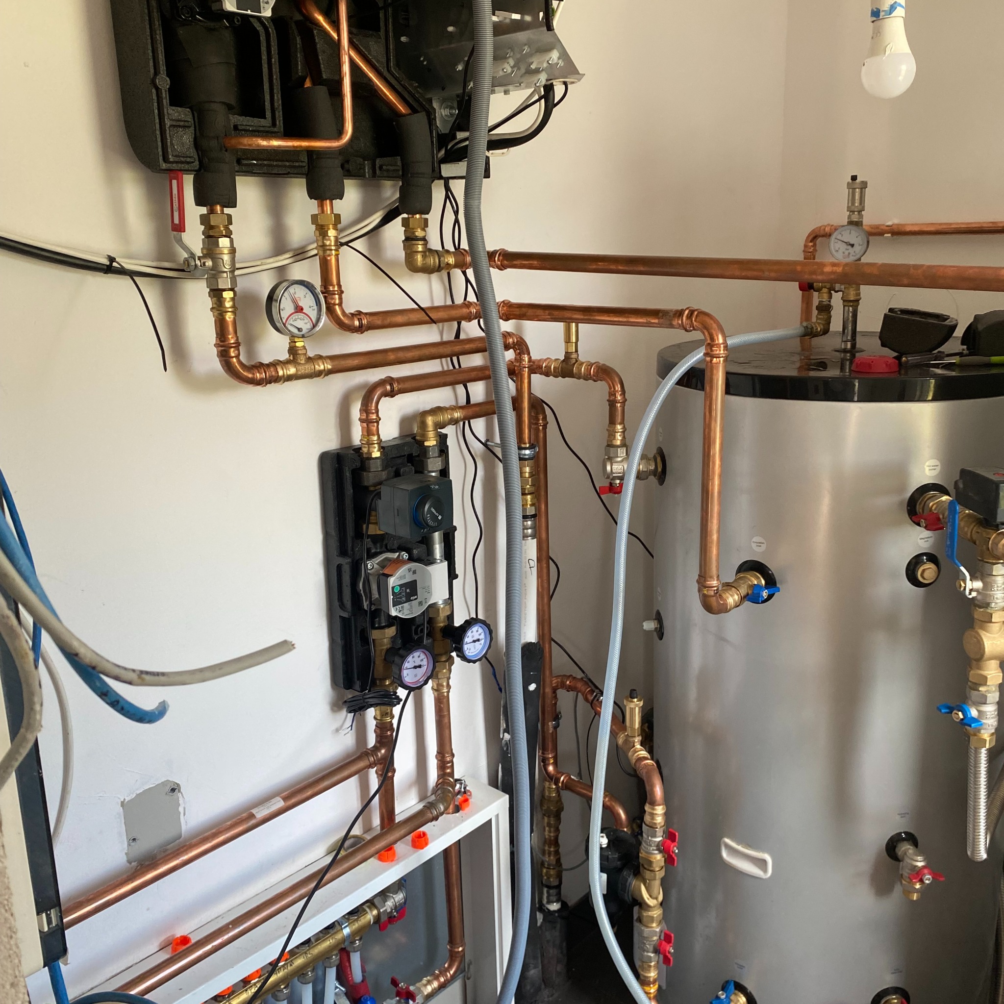 Instalacja grzewcza z miedzianymi rurami, zbiornikiem i czarną skrzynką rozdzielczą na białej ścianie. Widoczne manometry, zawory i kable. Żarówka zwisa z sufitu.