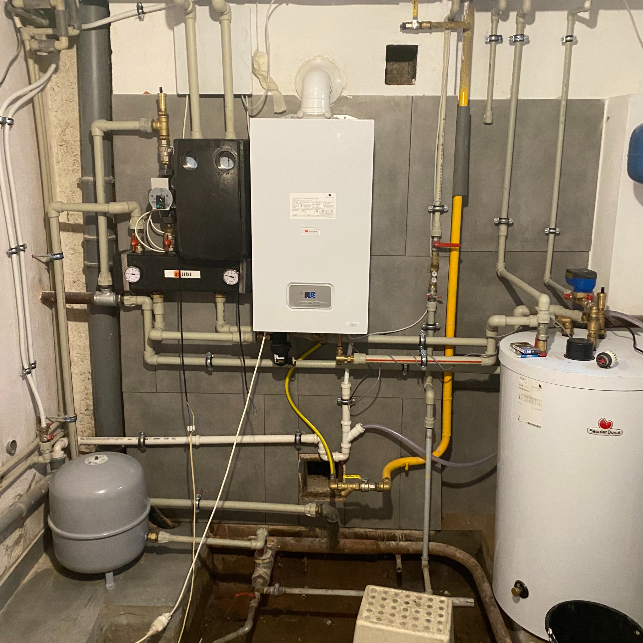 Złożona instalacja kotła gazowego z licznymi rurami, zaworami i zbiornikiem, widoczne także elementy marki Saunier Duval, całość na tle szarej ściany z płytek.