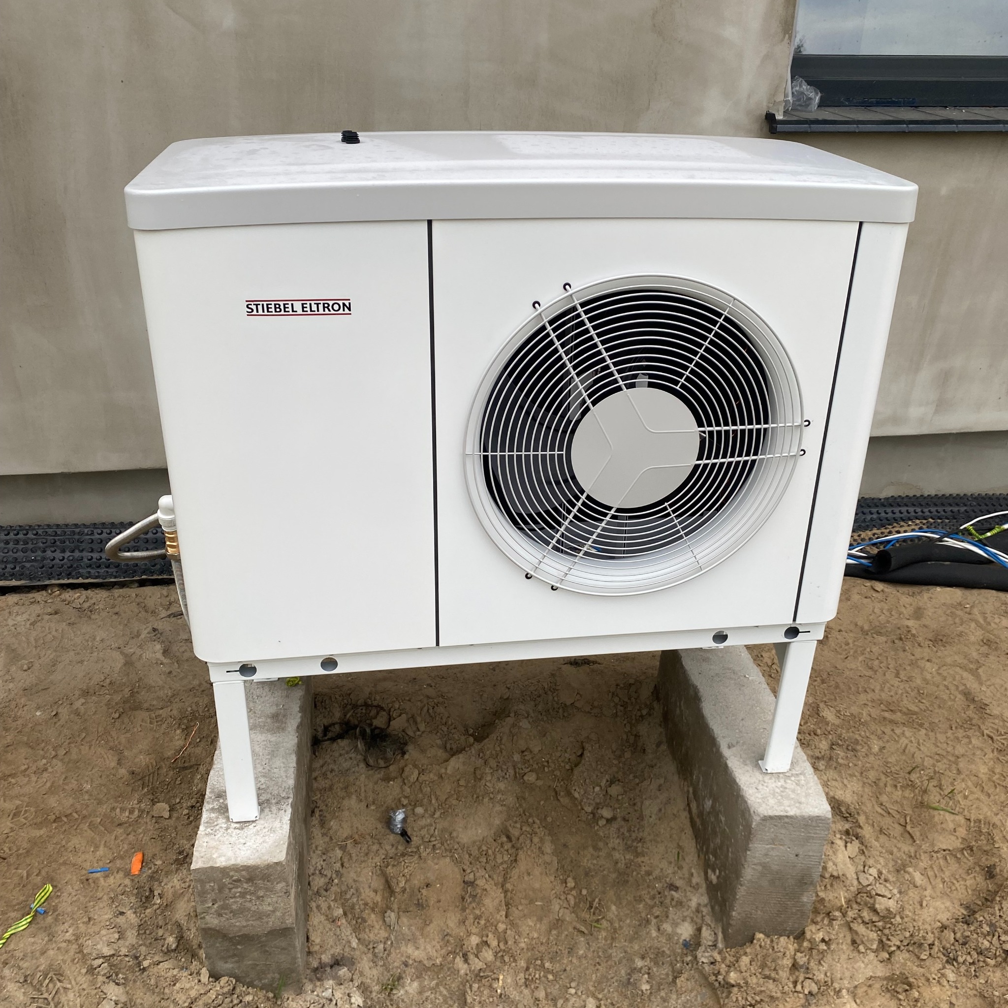 Biała pompa ciepła marki Stiebel Eltron zamontowana na betonowych podstawach przy ścianie budynku, widoczne podłączenia przewodów.