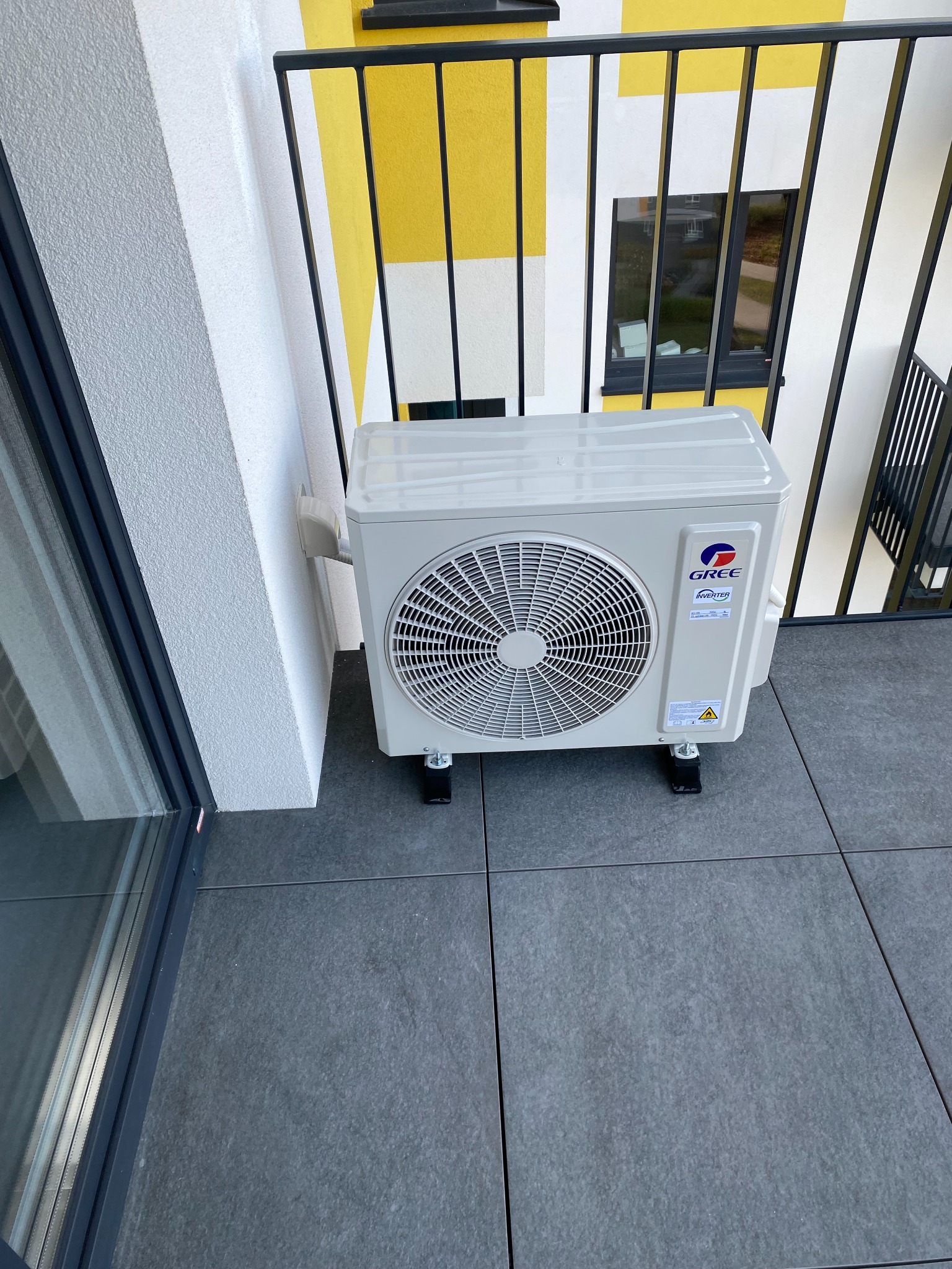 Biała jednostka zewnętrzna klimatyzacji marki Gree na balkonie z szarą podłogą z płytek, widoczna część budynku z żółto-białymi pasami i czarną balustradą.