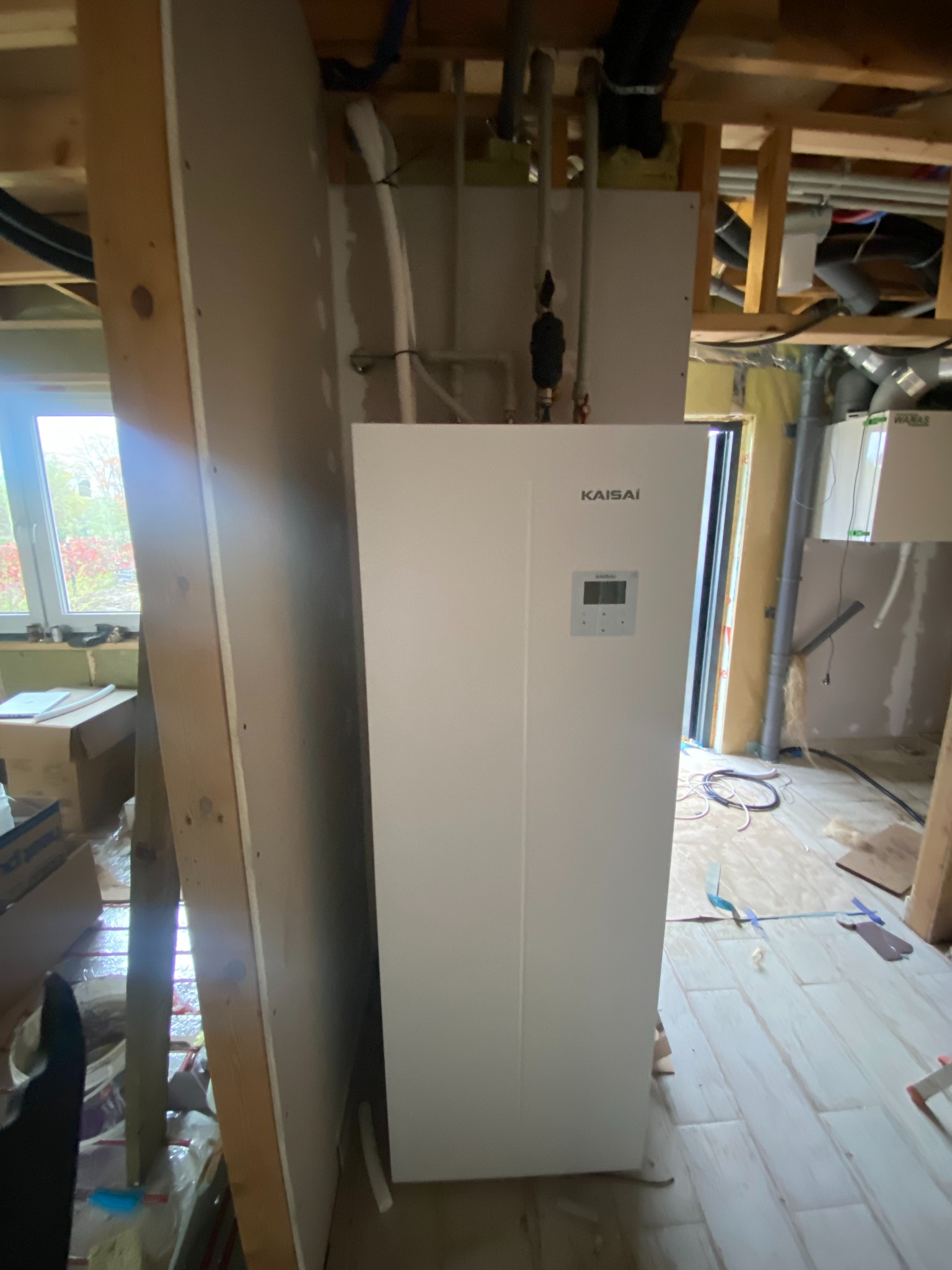 Biała pompa ciepła Kaisai zamontowana w surowym wnętrzu, widoczne rury instalacyjne i fragmenty konstrukcji budynku w trakcie wykończenia.