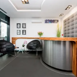 Nowoczesne wnętrze poczekalni z recepcją w klinice, z dwoma czarnymi fotelami o organicznym kształcie, szarą podłogą, drewnianą ladą recepcyjną z metalowym wykończeniem, dekoracyjną ścianą...