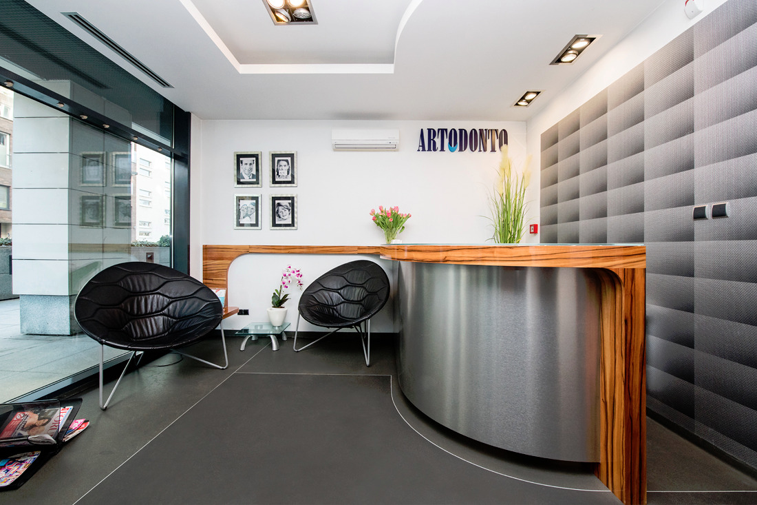 Nowoczesne wnętrze poczekalni z recepcją w klinice, z dwoma czarnymi fotelami o organicznym kształcie, szarą podłogą, drewnianą ladą recepcyjną z metalowym wykończeniem, dekoracyjną ścianą...