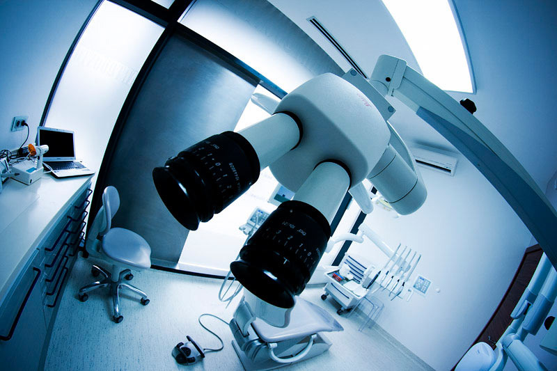 Nowoczesny mikroskop zabiegowy w chłodnym odcieniu błękitu, z widocznym pulpitem i fotelem dentystycznym w tle.
