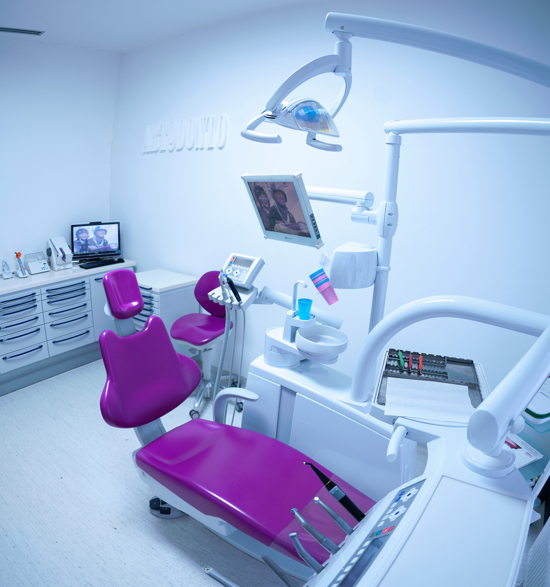 Nowoczesny gabinet z fioletowym fotelem dentystycznym, białym sprzętem i napisem 'Orthodonto' na ścianie, oświetlony chłodnym, niebieskawym światłem.