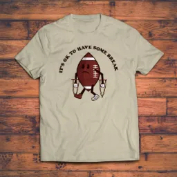 Beżowa koszulka z nadrukiem kreskówkowym przedstawiającym smutną piłkę do futbolu amerykańskiego na kulach, z napisem 'It's ok to have some break' na drewnianym tle.