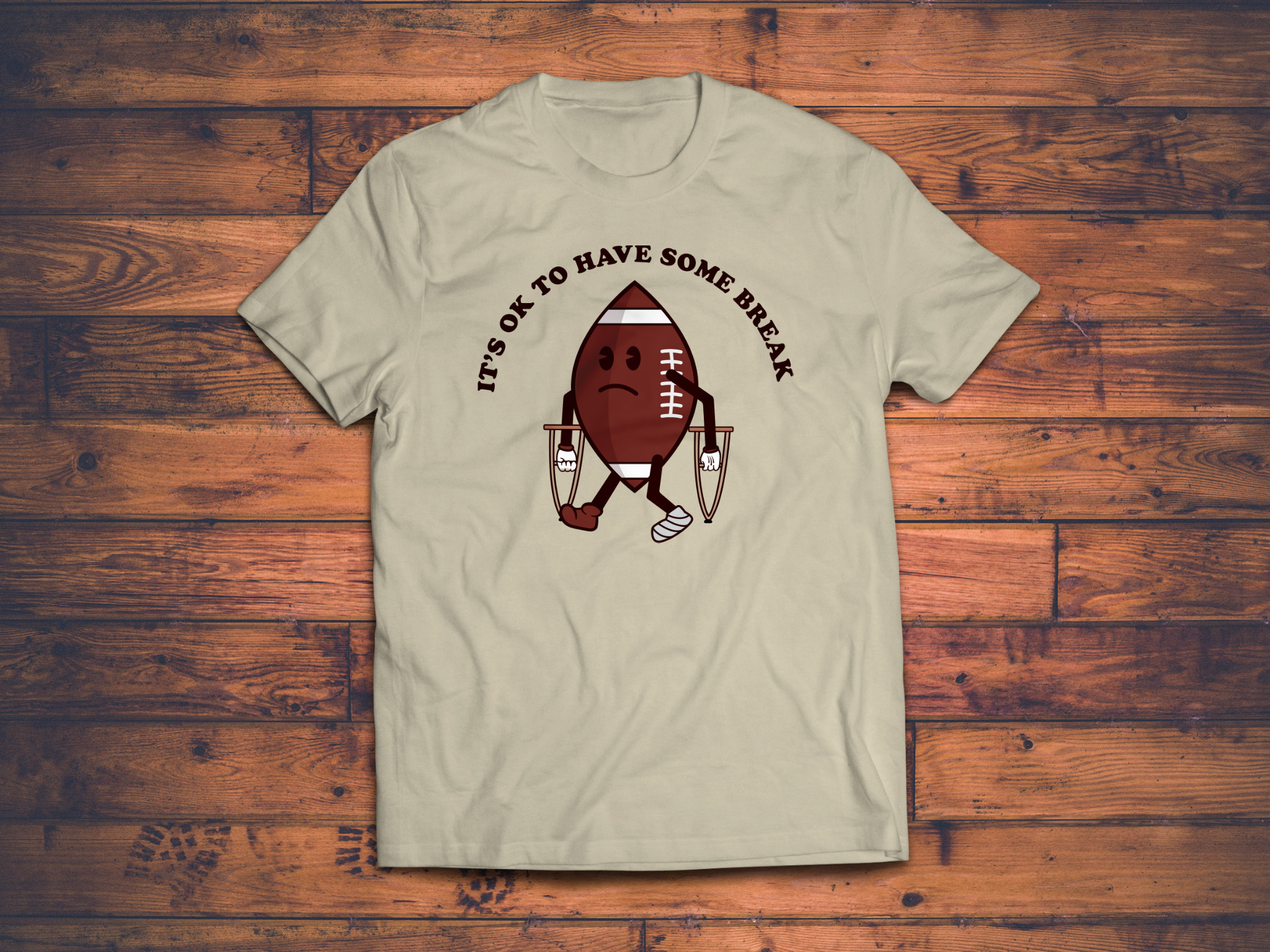 Beżowa koszulka z nadrukiem kreskówkowym przedstawiającym smutną piłkę do futbolu amerykańskiego na kulach, z napisem 'It's ok to have some break' na drewnianym tle.