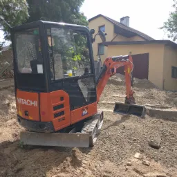 Pomarańczowa mini koparka Hitachi Zaxis 19 stoi na nierównym terenie z wykopaną ziemią, w tle budynek z beżową elewacją i zielone drzewa.