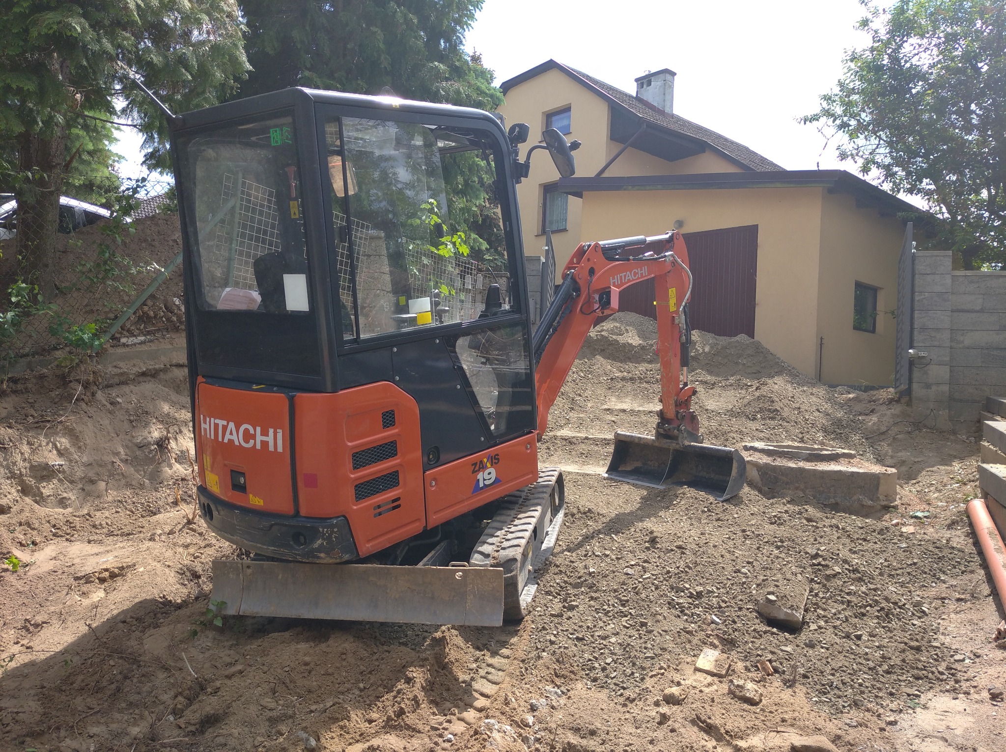 Pomarańczowa mini koparka Hitachi Zaxis 19 stoi na nierównym terenie z wykopaną ziemią, w tle budynek z beżową elewacją i zielone drzewa.