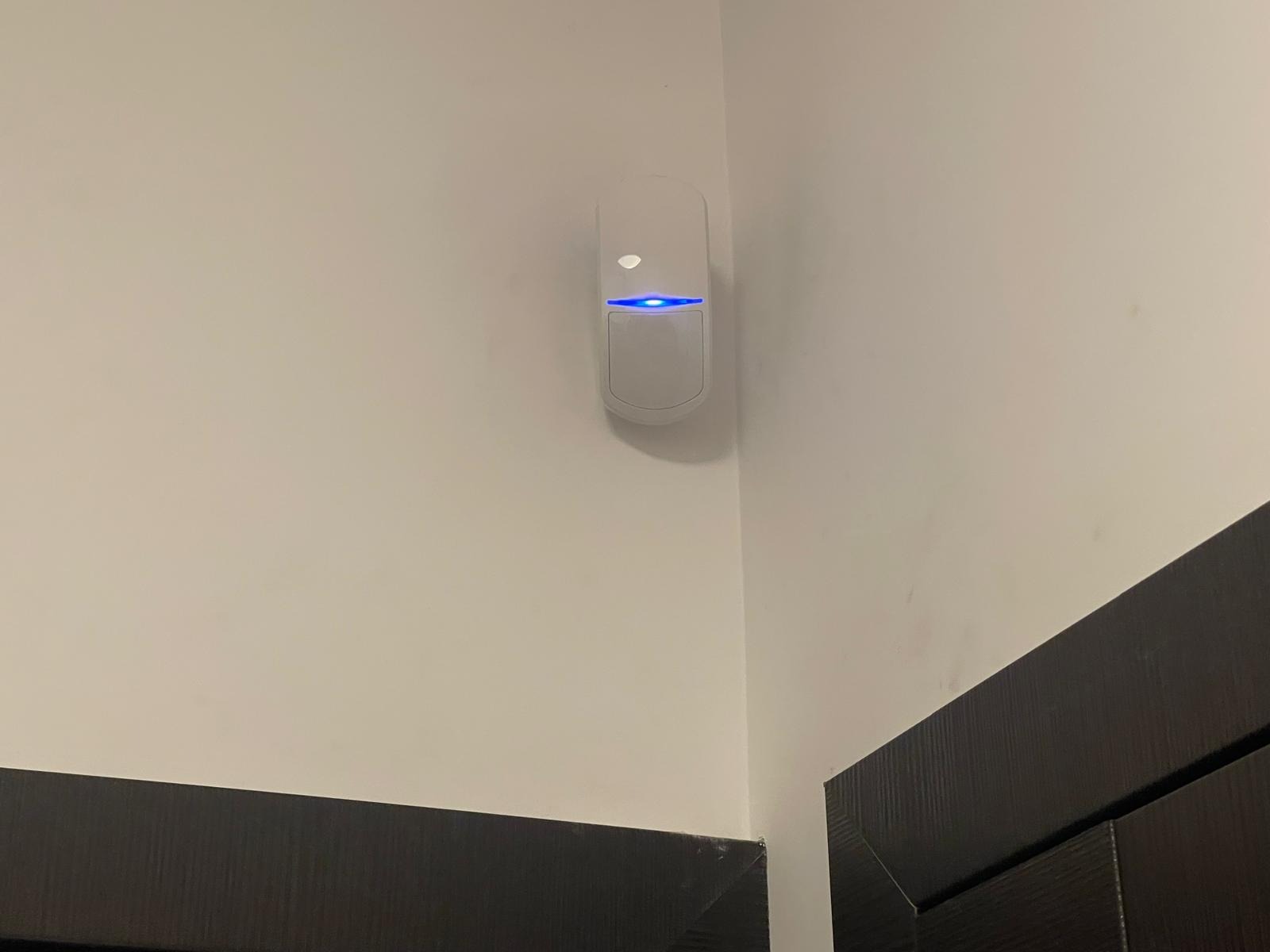 Zamontowana na ścianie czujka alarmowa z niebieską diodą, widoczny fragment futryny drzwi.