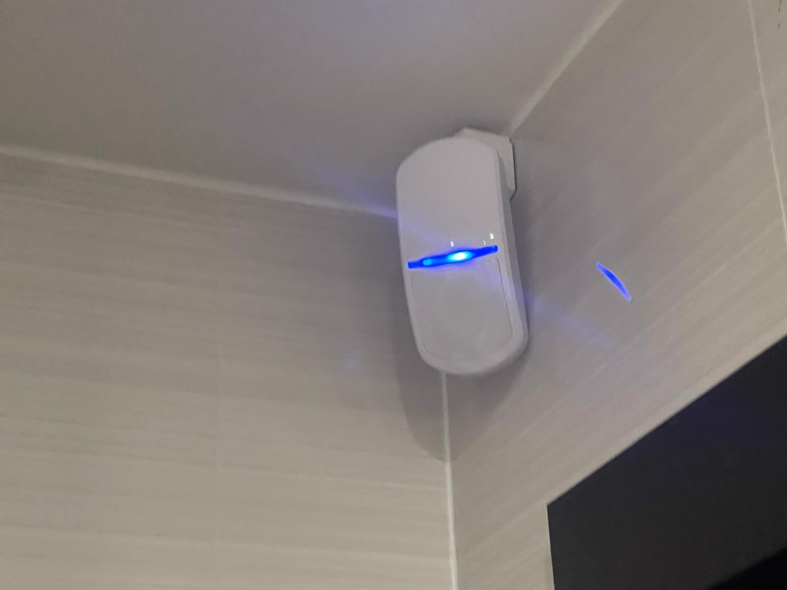 Biała czujka alarmowa z niebieską diodą LED zamontowana w rogu pomieszczenia, na ścianie wyłożonej płytkami imitującymi kamień.