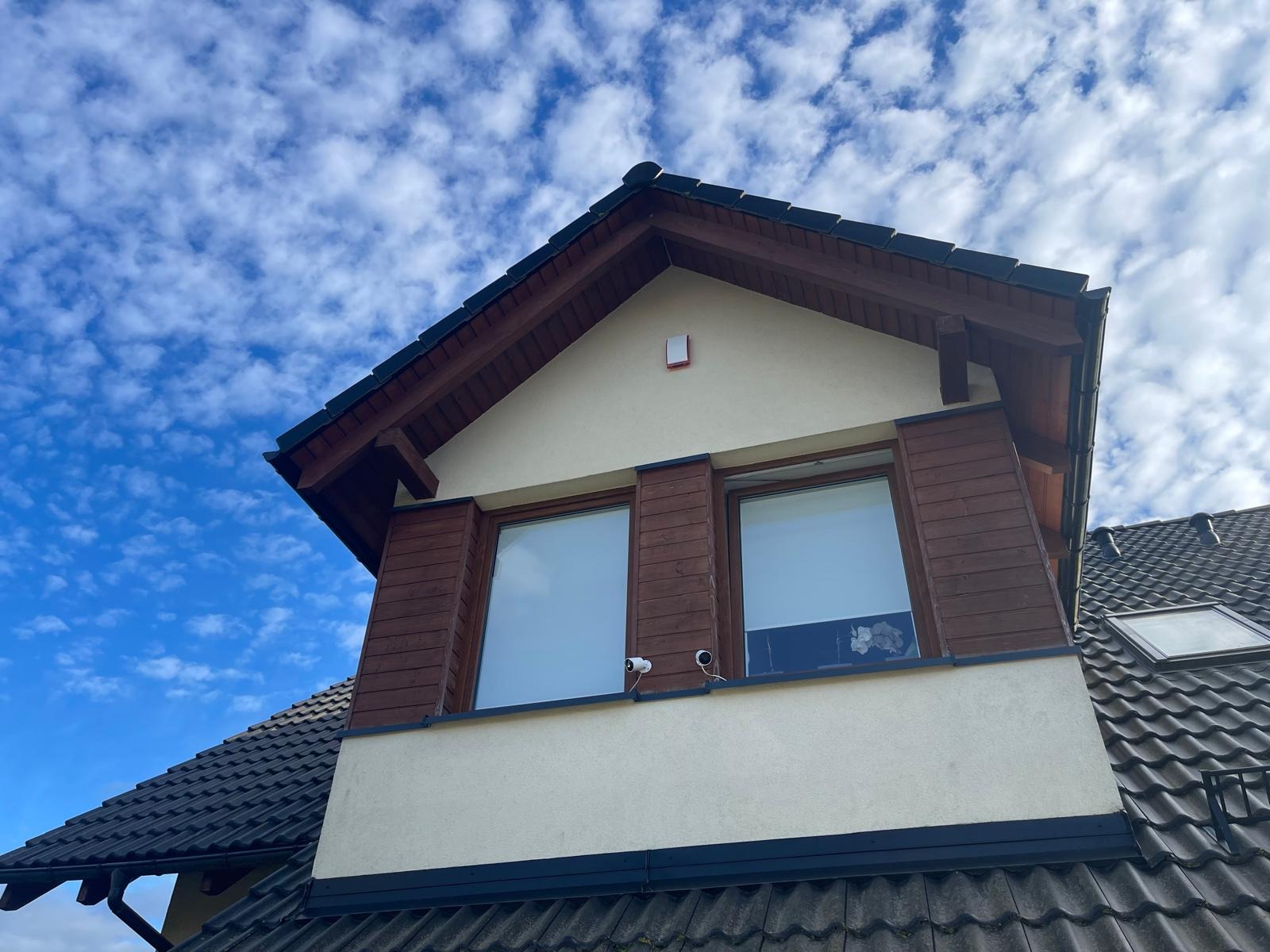 Dwie kamery monitoringu, zamontowane nad oknami na elewacji domu z drewnianą stolarką, na tle błękitnego nieba z chmurami.