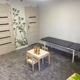 Gabinet logopedyczny z dwoma drewnianymi drzwiami z czarnymi paskami, tapetą z motywem roślinnym, szarym dywanem, kozetką z szarym obiciem, stoliczkiem z krzesełkami i zabawkami edukacyjnymi.