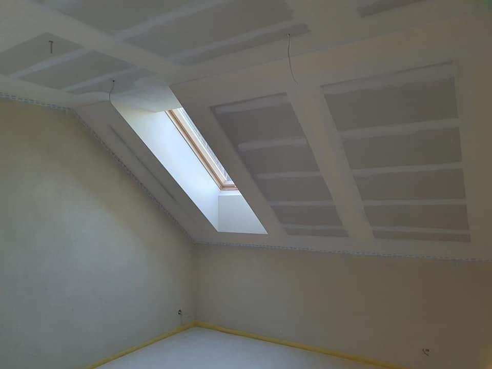 Poddasze z oknem dachowym w trakcie prac wykończeniowych; widoczne płyty na suficie i przygotowane ściany, zabezpieczona podłoga.