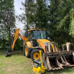 Żółta koparko-ładowarka JCB 860 i zagęszczarka gruntu Wacker Neuson na trawiastym terenie, w tle drzewa.