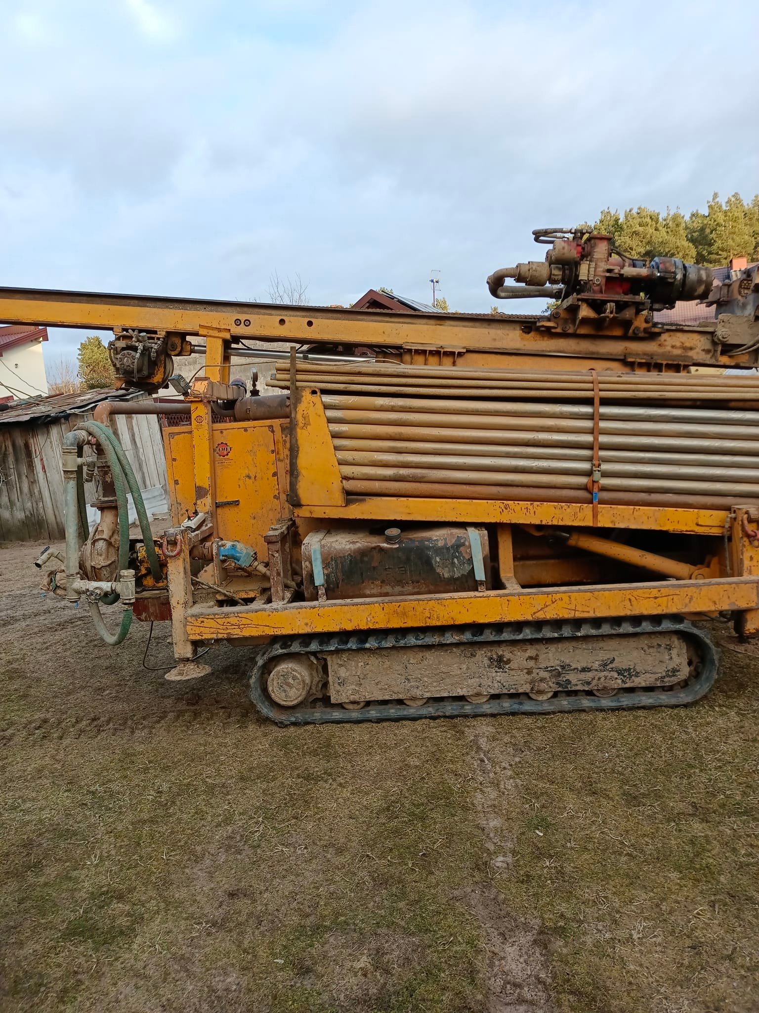 Żółta, gąsienicowa wiertnica do odwiertów pod pompy ciepła, widoczna w terenie trawiastym, z rurami wiertniczymi na górze i błotem na gąsienicach.